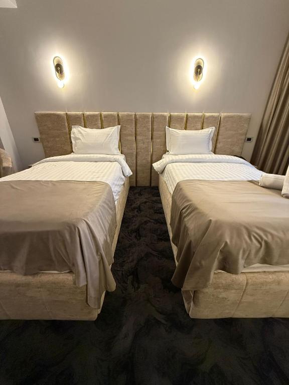 Două paturi twin simetrice cu tăblii capitonate crem și profil auriu, sub aplice de perete luminate.