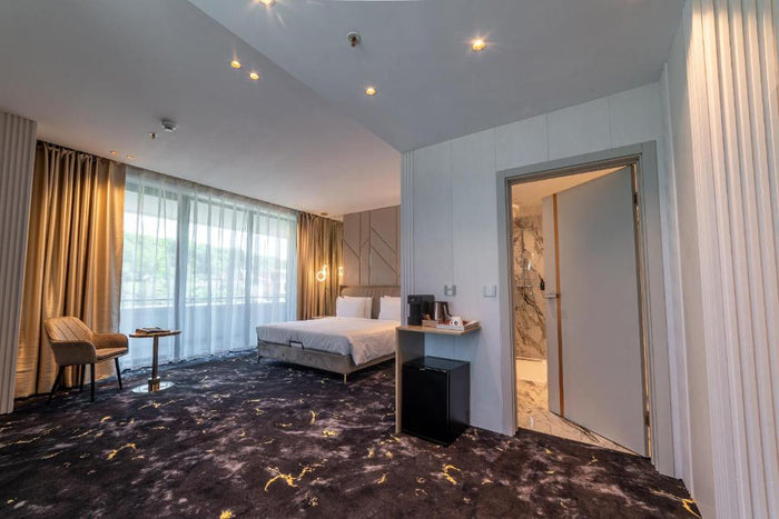 Cameră spațioasă cu minibar sub consolă, ușă deschisă spre baia placată cu marmură și mochetă bleumarin.