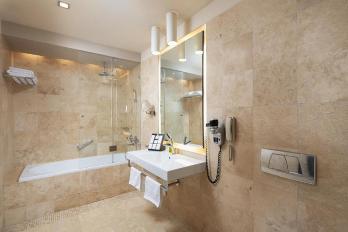 Baie placată cu travertin la Golden Tulip Cluj-Napoca, cu cadă cu paravan de sticlă, lavoar și oglindă iluminată.