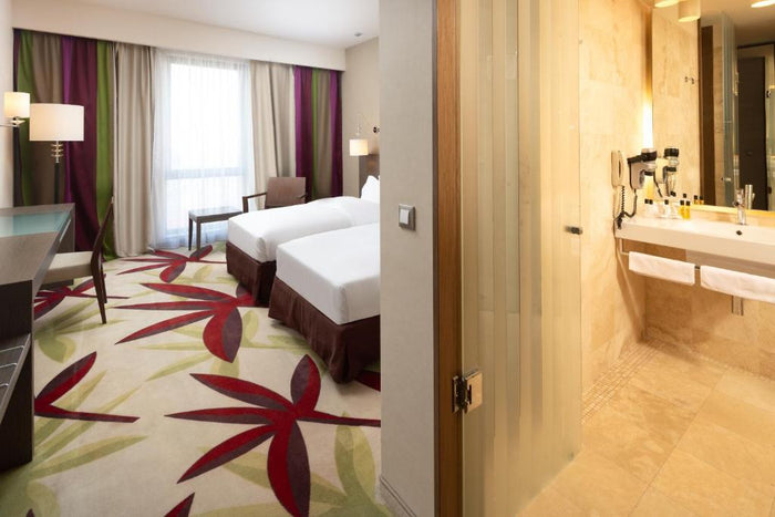 Cameră twin la Golden Tulip Cluj-Napoca, cu două paturi, birou și baie placată cu travertin văzută prin ușa deschisă.