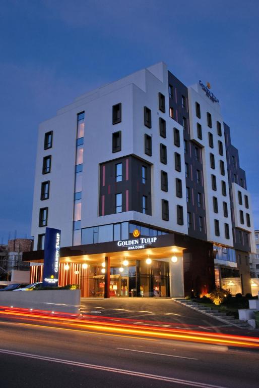 Fațada hotelului Golden Tulip Ana Dome iluminată seara, cu intrarea luminată cald și dâre de lumină pe bulevard.