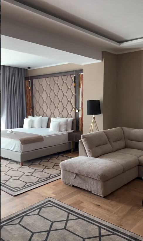 Apartament cu dormitor și zonă de living deschisă, canapea gri modulară și covoare geometrice pe parchet.