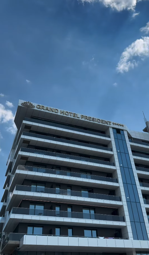 Fațada hotelului văzută de jos ziua, cu firma luminoasă pe acoperiș și balcoane etajate pe cer senin.