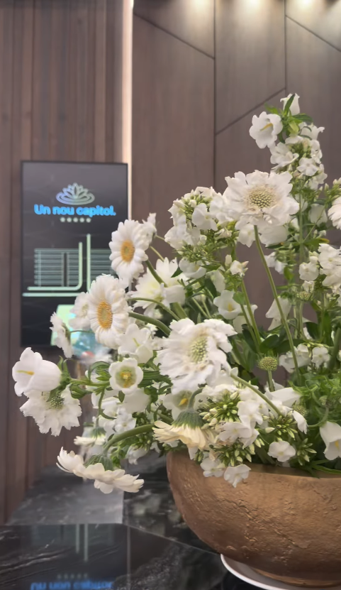 Aranjament floral cu flori albe într-un vas din lut, în holul cu pereți din lemn și ecran digital în fundal.