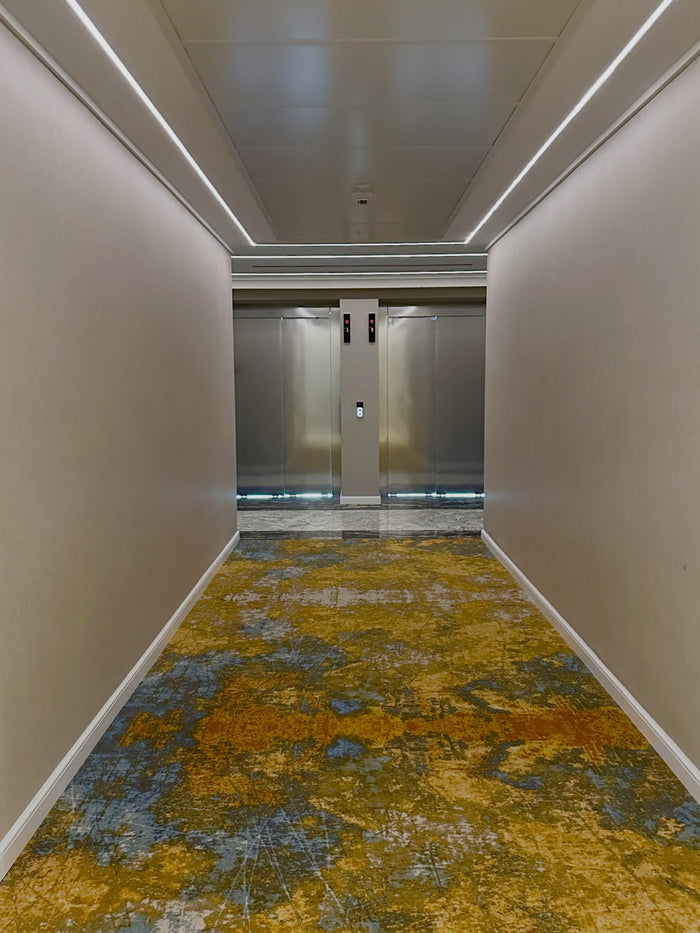 Hol de lift cu covor în nuanțe de auriu și albastru, uși de lift din inox și iluminat liniar pe tavan.