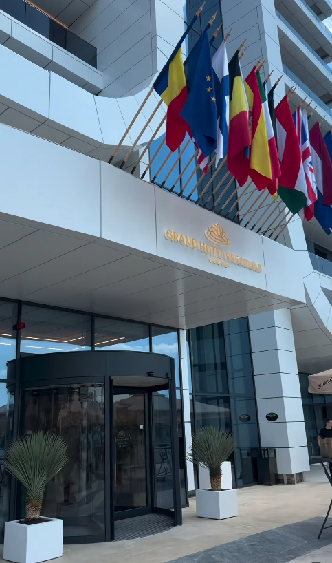 Intrarea principală cu ușă rotativă din sticlă, ghivece cu palmieri și drapele europene deasupra firmei.