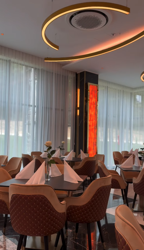 Mese de restaurant aranjate cu șervete împăturite triunghiular și trandafiri albi, sub un lustru auriu curbat.