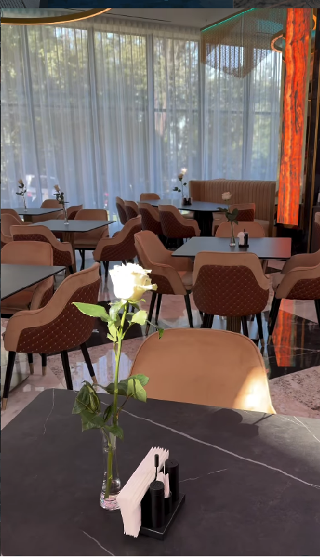 Sală de restaurant cu perdele transparente, scaune capitonate maro și un trandafir alb pe fiecare masă.