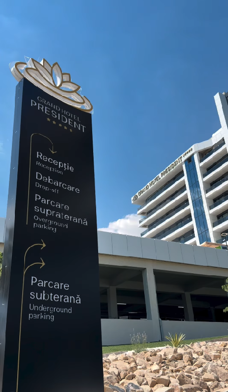 Totem indicator exterior cu sigla hotelului și direcții spre recepție și parcare, cu clădirea în fundal.