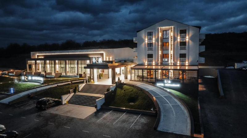 Clădirea hotelului luminată noaptea, cu firma Hotel Restaurant deasupra intrării și parcarea din față.