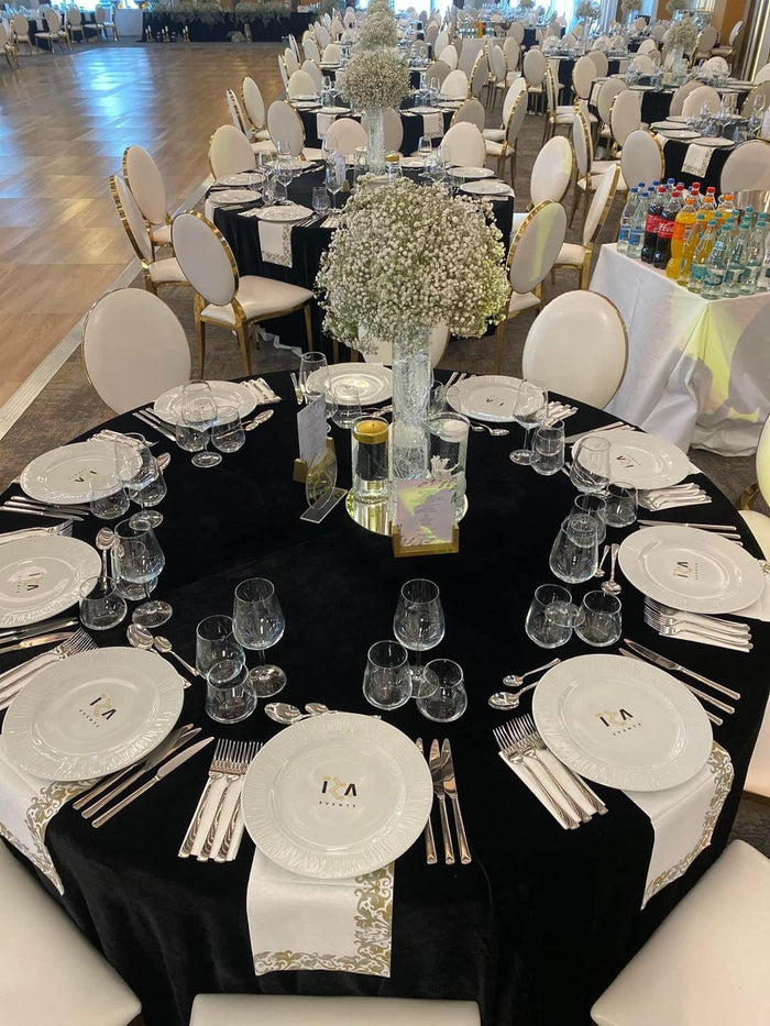 Masă de eveniment cu traverse albe brodate cu auriu și farfurii personalizate, cu ringul de dans în fundal.