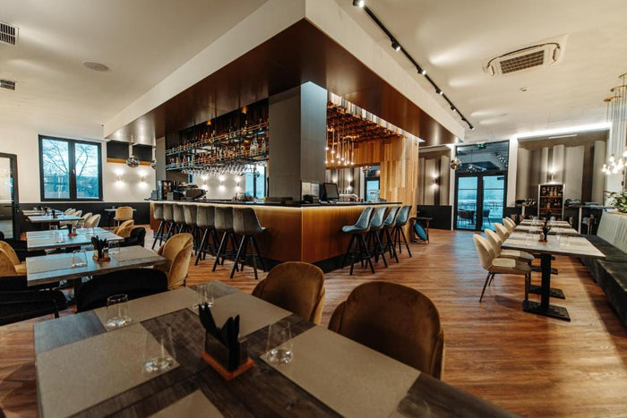 Sala restaurantului cu mese din lemn, scaune din catifea muștar și negre, iar în fundal barul cu taburete.