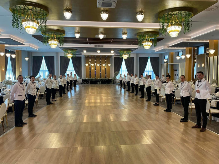 Sală de evenimente pregătită, cu ringul de dans liber și echipa de servire aliniată în ținute albe și negre.
