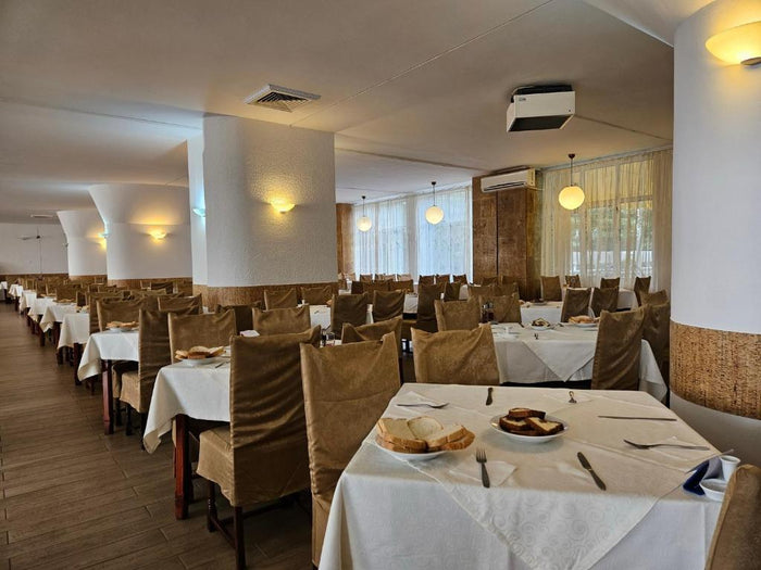 Sala de mese a Hotel Mureș, cu fețe de masă albe, huse maro pe scaune și aplice pe coloanele albe.