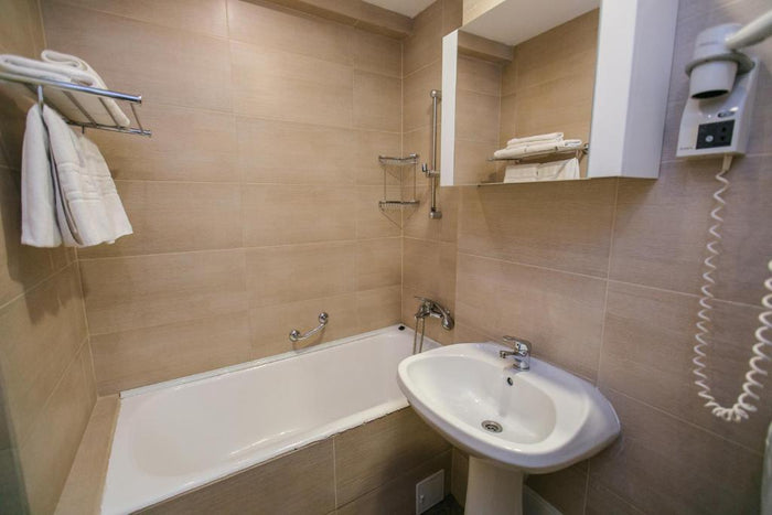 Baie la Hotel Nufărul, placată cu gresie bej, cu cadă, lavoar pe piedestal, uscător de păr și prosoape albe.