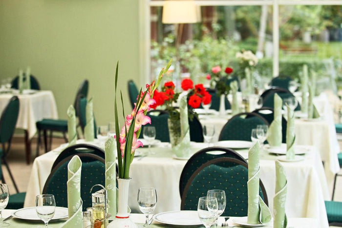 Mese aranjate la restaurantul Hotel Nufărul, cu șervete verzi împăturite, pahare de vin și gladiole roz în vază.