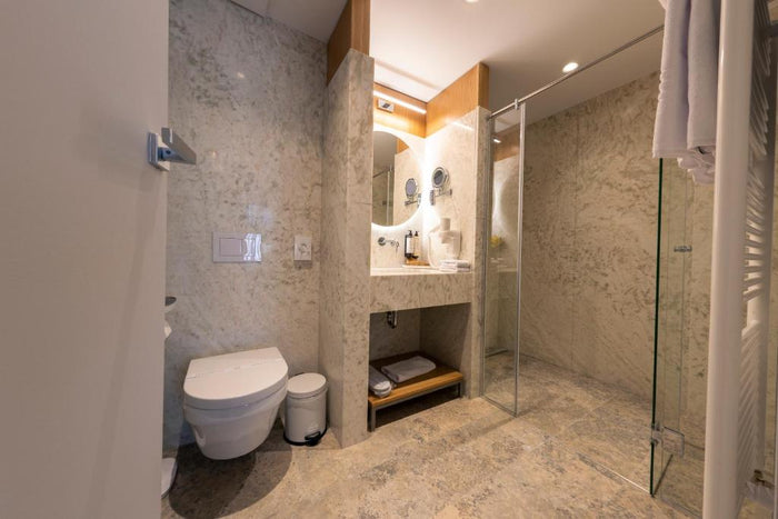 Baie placată cu marmură la Hotel One, cu cabină de duș din sticlă, oglindă rotundă și prosoape albe.