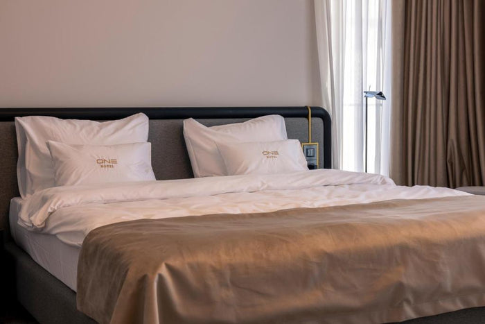 Pat dublu la Hotel One cu tăblie din piele neagră, perne brodate ONE Hotel și draperii bej la fereastră.