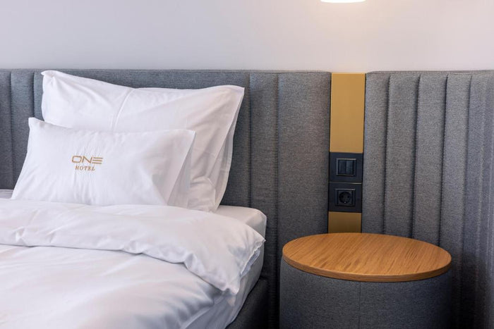 Noptieră rotundă din lemn lângă pat la Hotel One, cu panou capitonat gri și prize integrate.