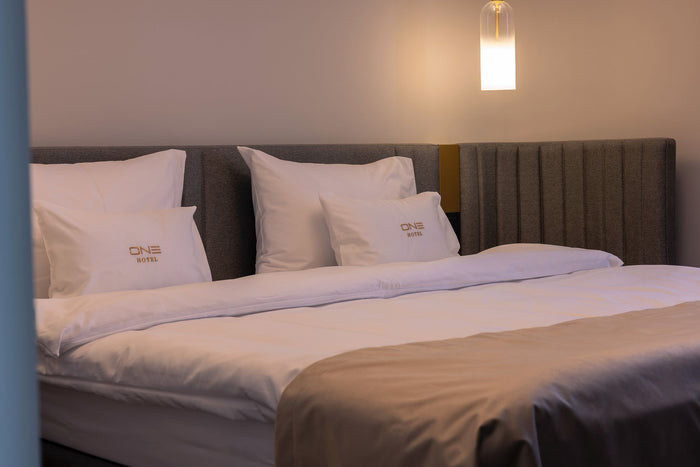 Pat dublu la Hotel One cu lenjerie albă și perne brodate cu sigla ONE Hotel, sub o lampă suspendată.