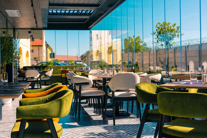 Terasa acoperită a restaurantului Hotel One, cu pereți din sticlă, scaune tapițate verde și mese din lemn.