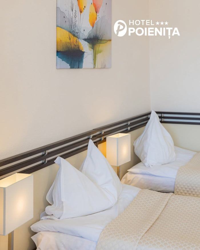 Detaliu cu două paturi la Hotel Poienița: perne albe așezate în vârf, cuverturi crem și veioze aprinse pe tăblie.