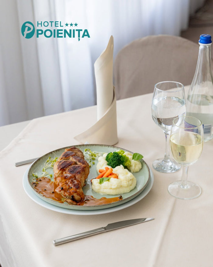 Preparat servit la restaurantul Hotel Poienița: friptură cu sos, piure și legume, alături de pahare de vin și apă.