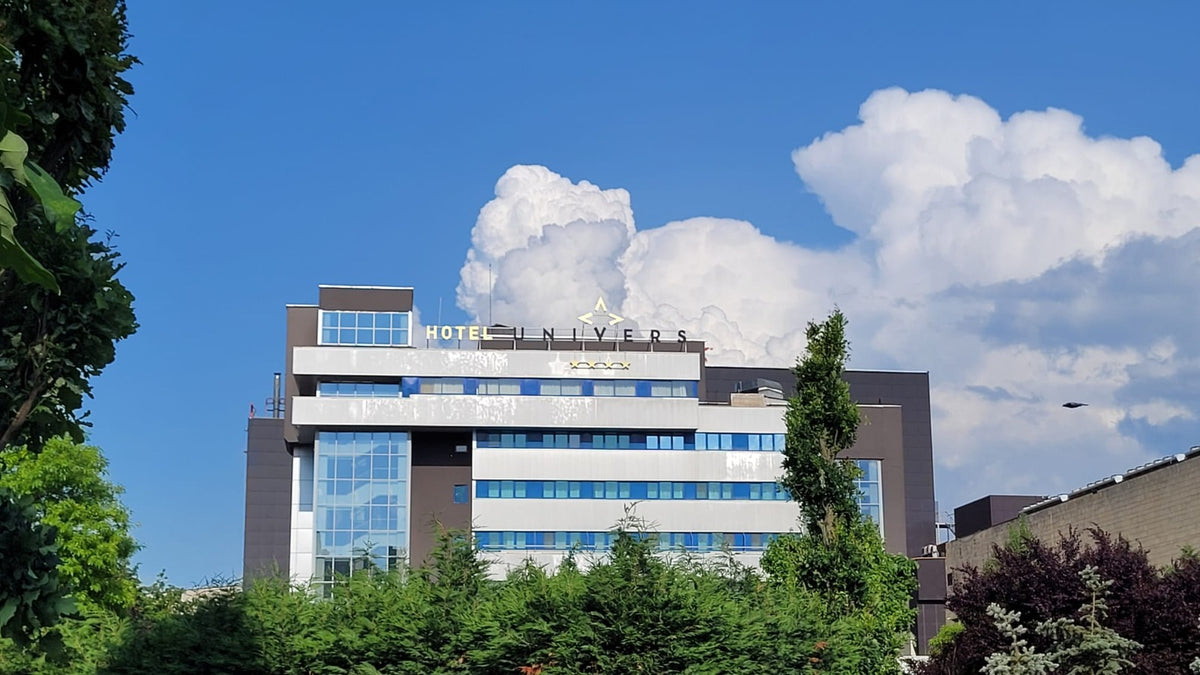 Sigla luminoasă HOTEL UNIVERS cu patru stele, montată pe aticul clădirii, pe fundal de cer albastru cu nori.