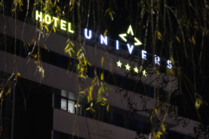 Firma luminoasă HOTEL UNIVERS aprinsă noaptea pe fațada clădirii, privită printre ramurile unei sălcii.