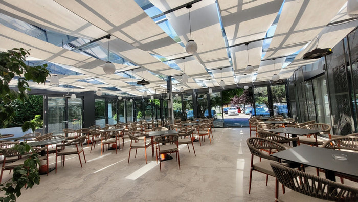 Terasă-restaurant vitrată, cu copertine textile sub luminator, fotolii din fibră împletită și mese negre.