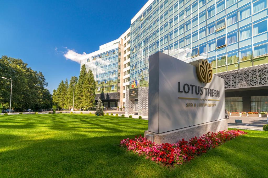 Fațada Lotus Therm Spa & Luxury Resort, cu sigla pe soclu din piatră, flori roșii și gazon îngrijit în prim-plan.