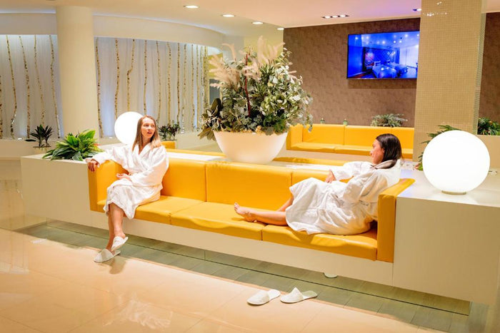 Două persoane în halate albe relaxându-se pe o canapea galbenă în lounge-ul SPA, cu papuci lăsați pe podea.