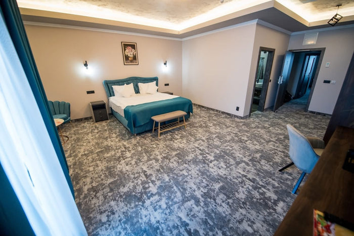 Cameră matrimonială văzută de la intrare, cu banchetă la picioarele patului, mochetă gri și ușa băii deschisă.