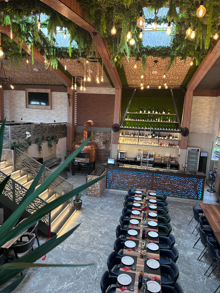 Restaurantul văzut de la etaj, cu rafturi de sticle pe perete vegetal, masă lungă pregătită și scara iluminată.