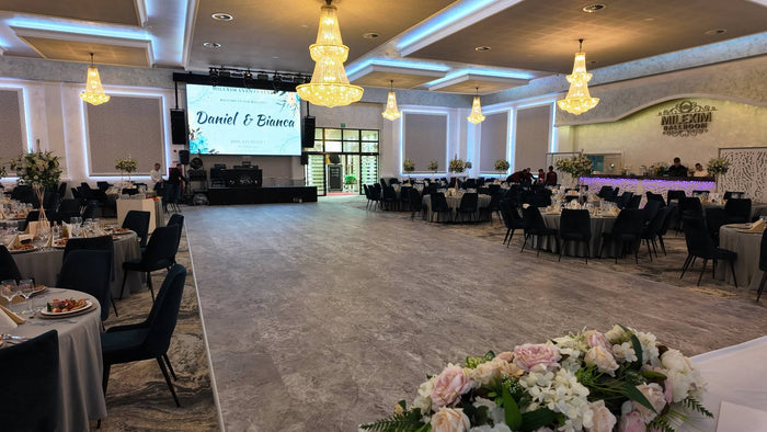 Sală de evenimente cu ecran de proiecție pe scenă, candelabre aurii, mese rotunde pregătite și bar în fundal.