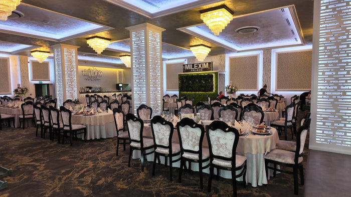 Sala Milexim Palace cu mese lungi, scaune baroc negre cu tapițerie florală, candelabre aurii și perete vegetal.