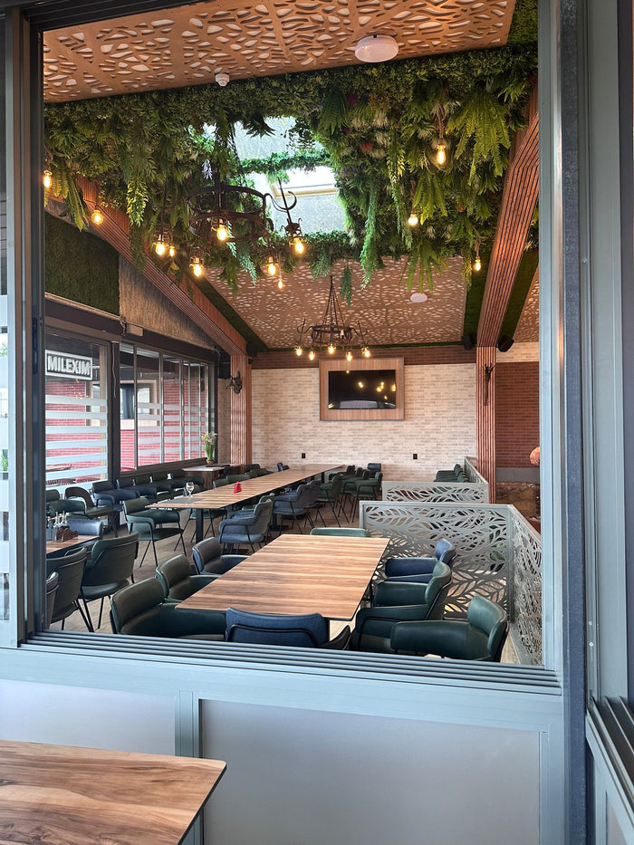 Sala restaurantului privită prin fereastra glisantă, cu plafon vegetal, candelabre din fier forjat și mese lungi.