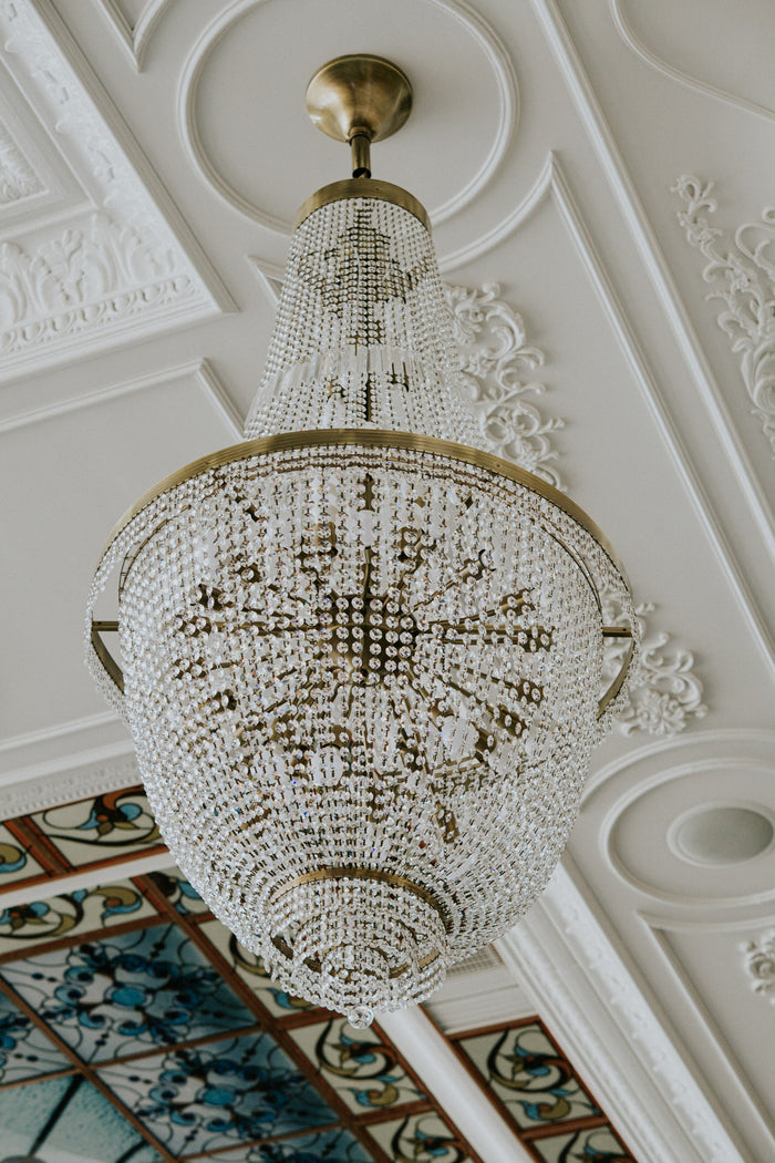 Candelabru din cristal cu structură aurie, suspendat de un tavan alb ornamentat cu stucaturi florale.