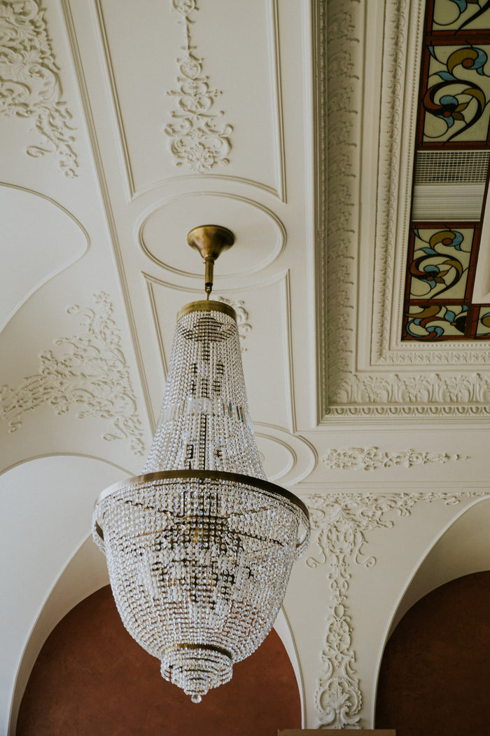 Candelabru din cristal în formă de coș, fotografiat de jos, lângă tavanul stucat și un panou de vitraliu.