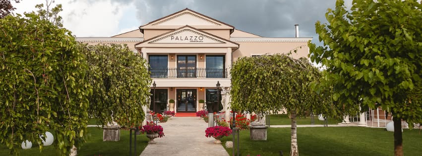 Fațada Palazzo Wedding Hall la amurg, cu alee spre intrare flancată de copaci și lumini decorative calde.