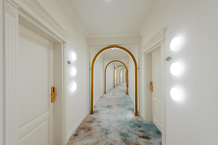 Hol de etaj cu arcade aurii succesive, uși albe cu numere de cameră, aplice rotunde și mochetă în tonuri de bleu.