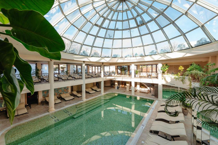 Piscină interioară sub o cupolă din sticlă, cu șezlonguri albe pe margine și plante verzi de jur împrejur.