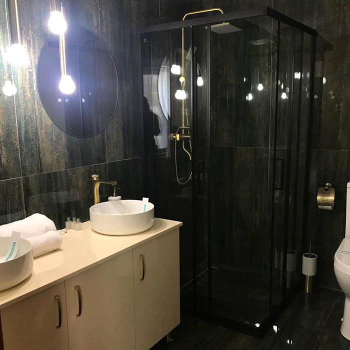 Baie cu lavoar tip bol, baterie aurie, oglindă rotundă și cabină de duș cu profile negre și duș fix auriu.