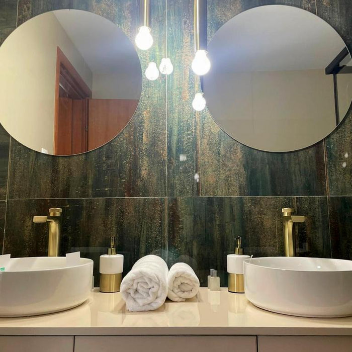 Baie cu două lavoare tip bol, baterii aurii, oglinzi rotunde și prosoape albe rulate, pe faianță închisă.