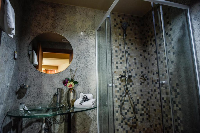 Baie cu lavoar din sticlă, oglindă rotundă și cabină de duș placată cu mozaic, cu prosoape și uscător de păr.
