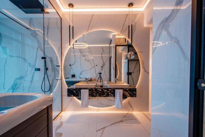 Detaliu al blatului cu două lavoare roz pudră sub o oglindă rotundă retroiluminată, cu oglindă cosmetică alăturată.