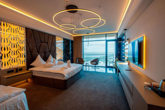 Cameră spațioasă cu tăblie maro cu model geometric, candelabru din inele suspendate și fereastră panoramică spre balcon.