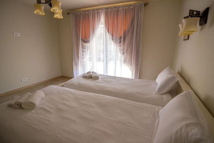 Cameră cu două paturi twin, prosoape rulate pe cuverturi și draperii albe cu accent portocaliu la fereastră.