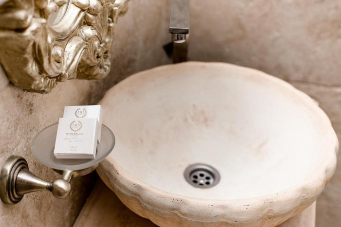 Lavoar din piatră naturală cu robinet modern din inox, cu săpunuri personalizate Wonderland pe un suport rotund alăturat.
