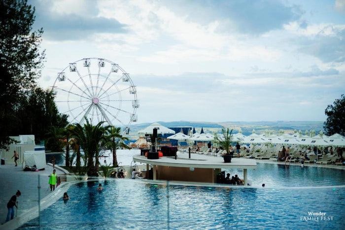 Piscina exterioară a Wonderland Cluj Resort, cu roata panoramică, șezlonguri și umbrele albe pe margine.